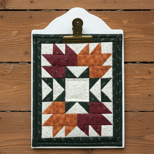 A quilted fabric wall hanging or plaque with a geometric starburst pattern, mounted on a white board and clipped at the top, displayed against a rustic wooden plank background.