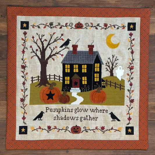 A square cross-stitch or needlepoint quilt featuring a Halloween scene with a black haunted house, orange pumpkins, bare trees, crows, a ghost, a yellow moon, and the text 'Pumpkins glow where shadows gather' on a wood background.