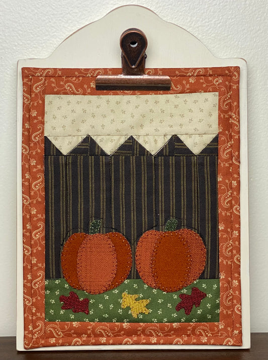 Wooden display board holding one of the clipboard quilts with pumpkins and leaves on a white background