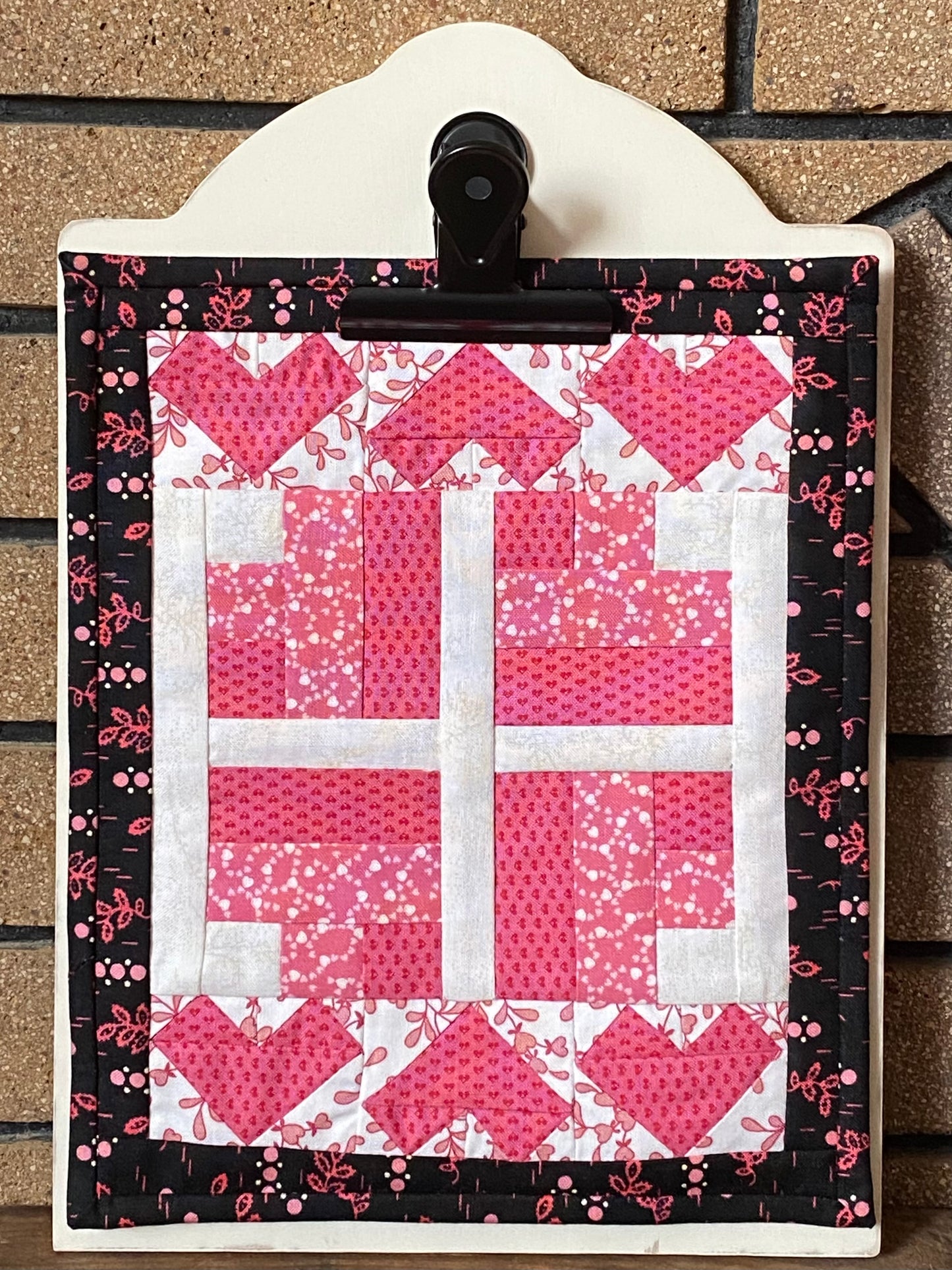 Wooden display board with a black clip holding one of the clipboard quilts with hearts, leaning against a brick wall.