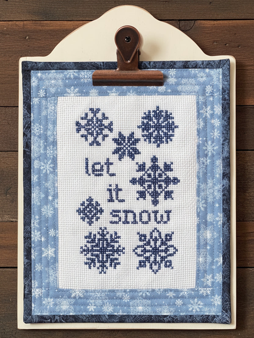 Cross-stitched panel with snowflakes and 'let it snow' text framed with a blue fabric border, clipped to a wooden board.
