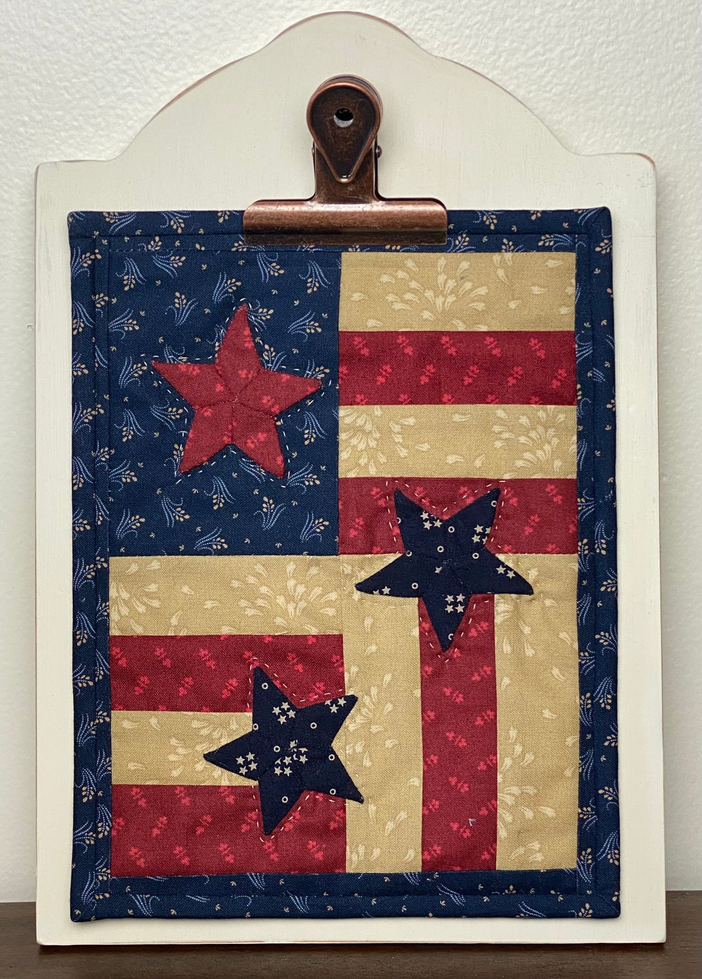 Wooden display board with bronze clip holding one of the clipboard quilts with flag and stars on a white background