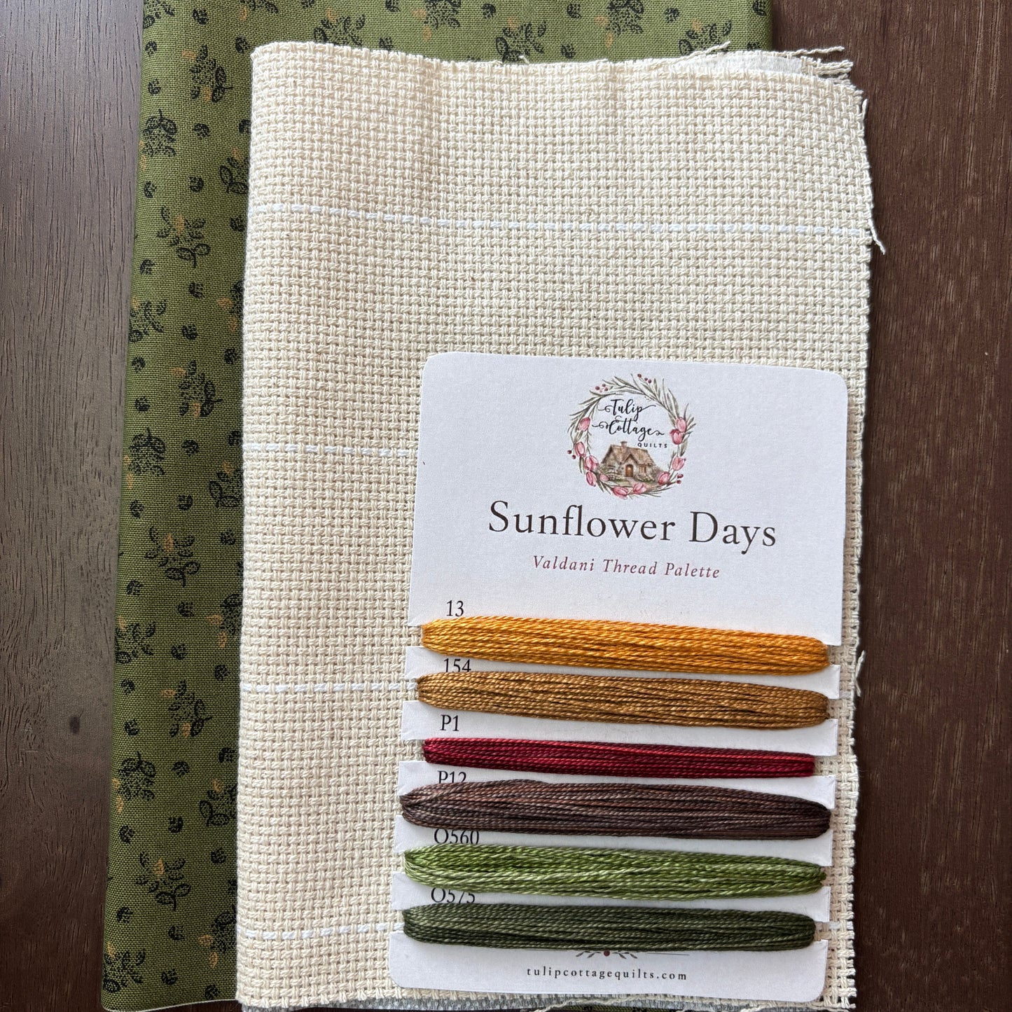 A fabric and thread palette set called 'Sunflower Days', featuring a beige card with six thread samples in yellow, gold, red, brown, sage green, and olive green, layered over a patterned olive green fabric, all displayed on a dark wood background.