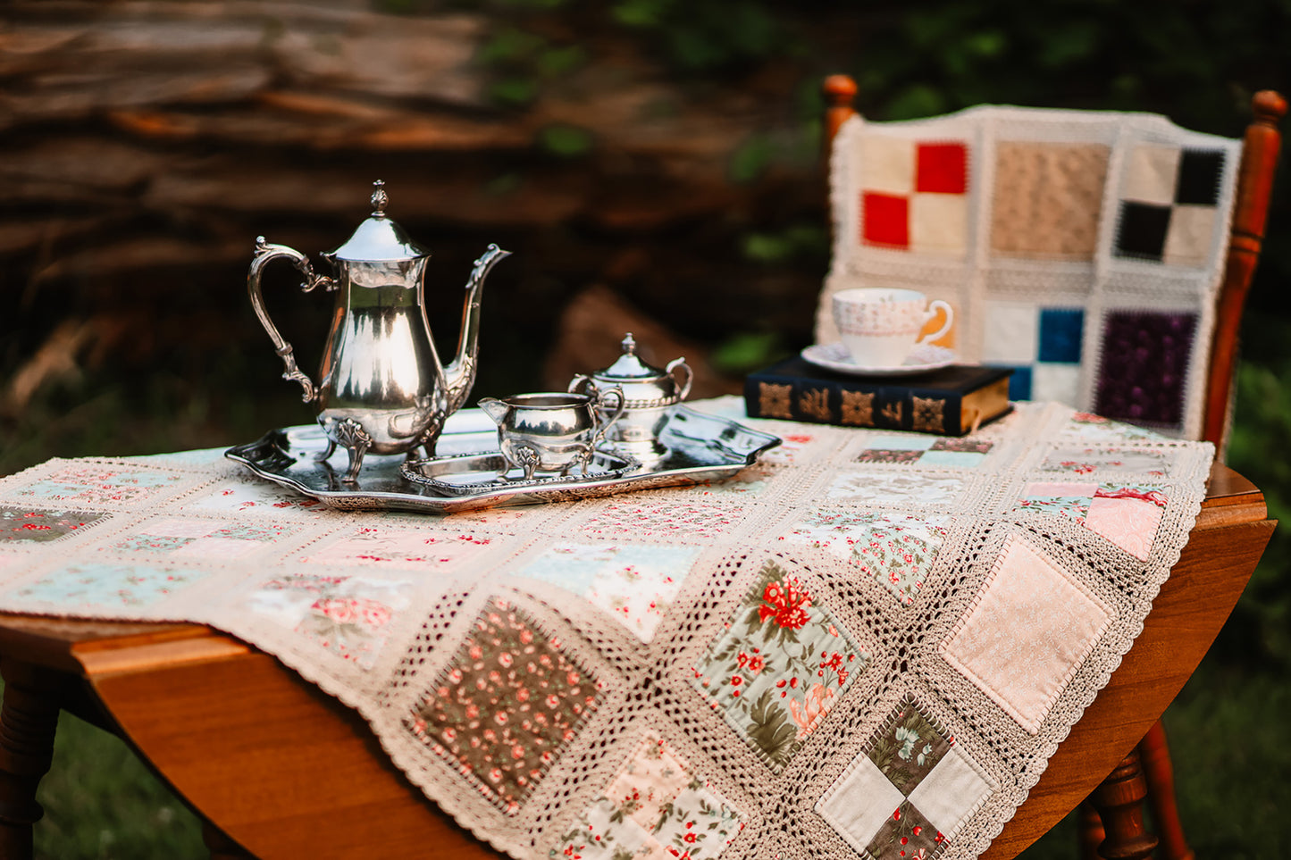 Silver teapot set and a crochet quilt set on a table with a chair in the background.