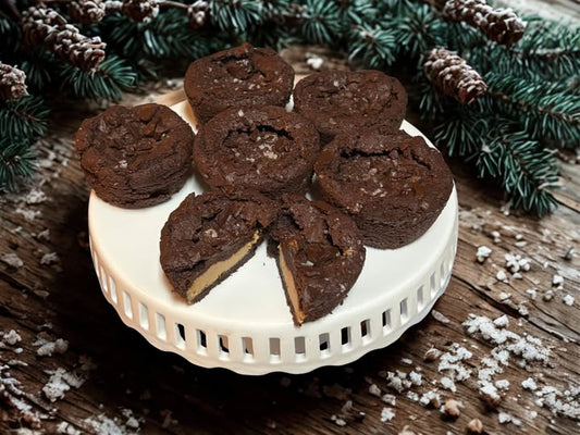 chocolate and peanut butter cookies on a cake stand sitting on a wooden surface with powdered sugar and pine boughs on the table around it.
