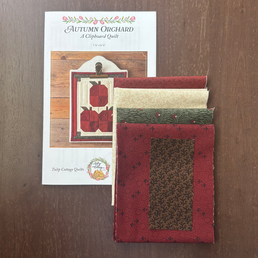 A folded quilt made from fabric samples is displayed next to its printed pattern booklet on a dark wood surface. The pattern is titled 'Autumn Orchard A Clipboard Quilt' by Tulip Cottage Quilt.