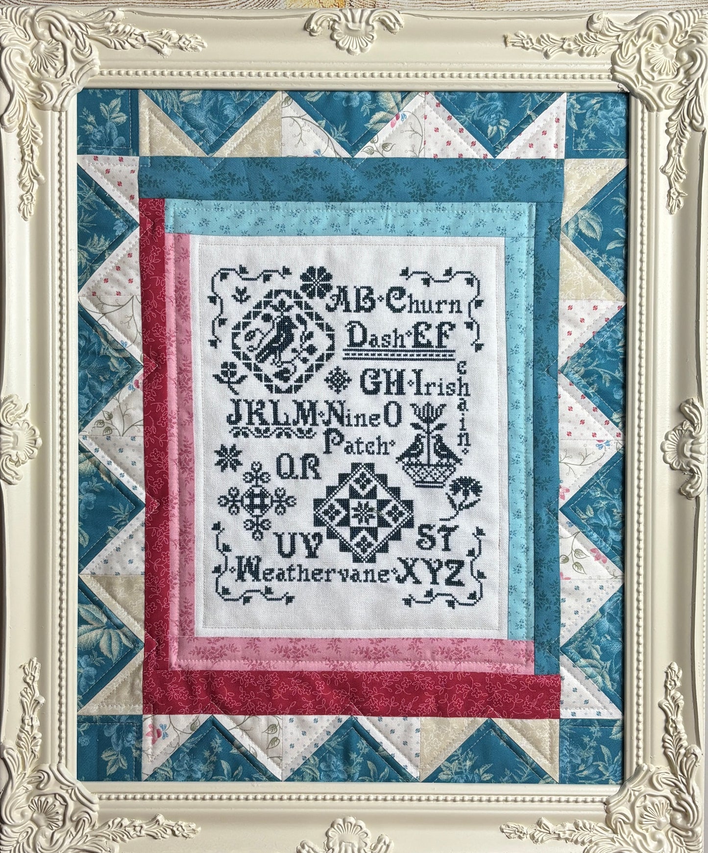 Quilt with cross-stitched text and decorative border in a decorative frame.