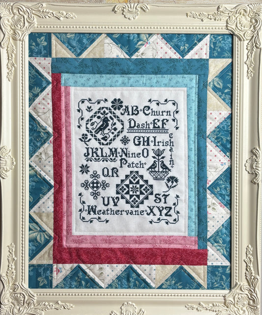 Quilt with cross-stitched text and decorative border in a decorative frame.