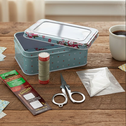 Sewing kit with floral-patterned tin, scissors, thread, needles, and a bag sitting on a wooden table with a cup of coffee.