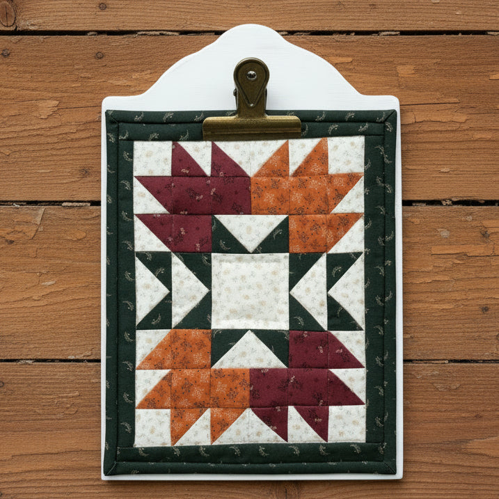 A quilted fabric wall hanging or plaque with a geometric starburst pattern, mounted on a white board and clipped at the top, displayed against a rustic wooden plank background.