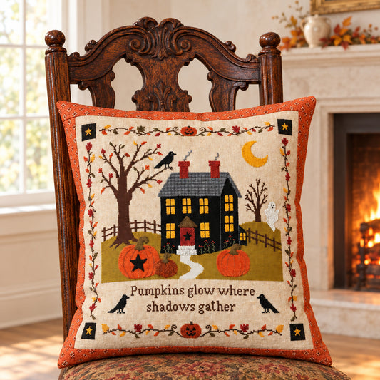 A square decorative throw pillow with an autumn Halloween design, displayed on a dark wooden chair inside a living room decorated with fall decor. The pillow features a black haunted house, pumpkins, crows, and the text 'Pumpkins glow where shadows gather'.
