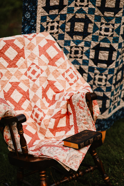 Quilt with geometric pattern draped over a wooden chair against a quilted backdrop.