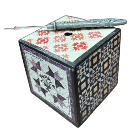 Decorative cube with geometric quilt patterns on a white background and seam ripper in the top.