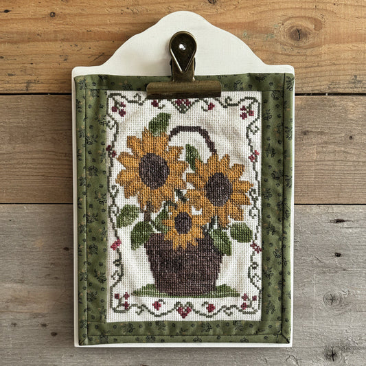 A rectangle cross-stitch sampler featuring a sunflower and basket design, displayed on a white clipboard with a metal clip. The sampler has an olive green patterned border and is set against a wooden background.