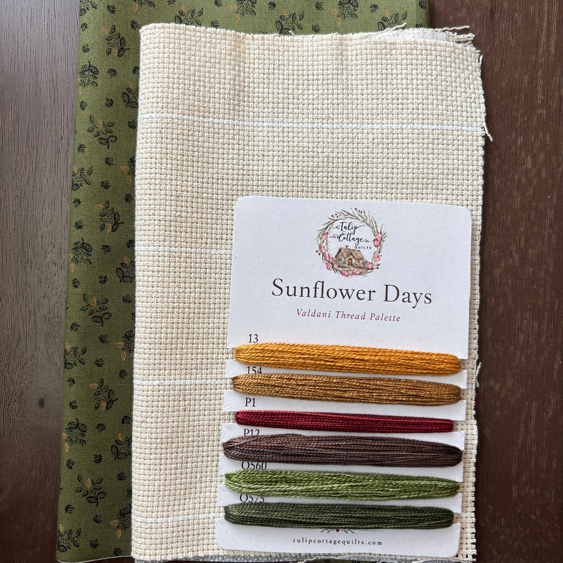 A fabric and thread palette set called 'Sunflower Days', featuring a beige card with six thread samples in yellow, gold, red, brown, sage green, and olive green, layered over a patterned olive green fabric, all displayed on a dark wood background.
