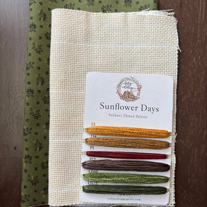 A fabric and thread palette set called 'Sunflower Days', featuring a beige card with six thread samples in yellow, gold, red, brown, sage green, and olive green, layered over a patterned olive green fabric, all displayed on a dark wood background.