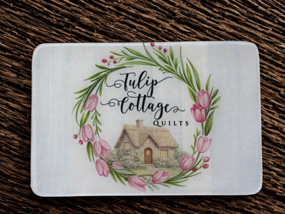 Card with 'Tulip Cottage Quilts' design featuring a cottage and floral wreath on a wooden surface.